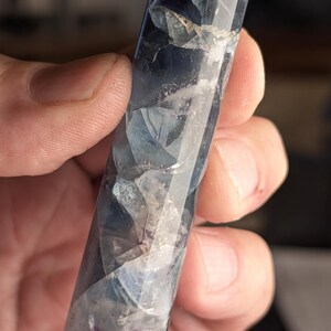 May include: A polished, elongated crystal with a rounded top. The crystal is primarily blue and white, with areas of purple and clear transparency. The facets catch the light, creating a shimmering effect. The crystal is held in a hand.