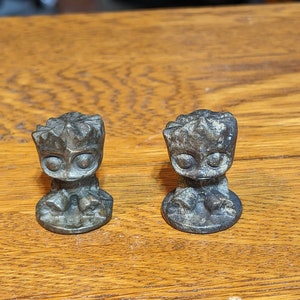 May include: Two small, gray, detailed figurines of a character from the Guardians of the Galaxy movie, Groot, standing on a wooden surface.