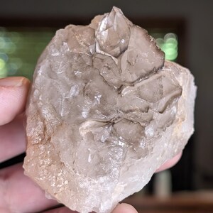 May include: A raw, natural quartz crystal cluster. The crystal features a mix of clear and smoky quartz, with a rough, textured surface and a prominent, pointed crystal formation at the top. The color is a mix of clear, white, and light brown.