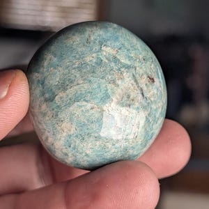 Puede incluir: Una piedra pulida y redonda con aspecto moteado. La piedra es principalmente de color verde azulado claro con parches blancos y marrones. La superficie tiene una textura ligeramente rugosa. La piedra se sostiene en una mano.