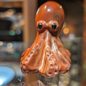 May include: A detailed, reddish-brown octopus figurine with black eyes. The sculpture has a textured surface with white and light brown veining. The octopus is displayed on a reflective surface, showcasing its intricate details.
