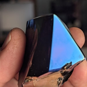 May include: A hand holding a triangular, iridescent crystal. The crystal has a dark, reflective surface with a vibrant blue and purple hue on one side. The edges appear rough and fractured.