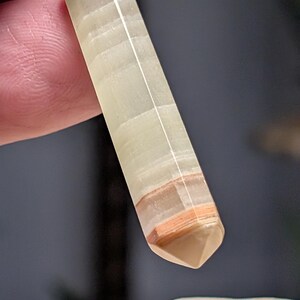 May include: A pendant featuring a long, hexagonal, pale green and translucent stone point with horizontal bands of white and reddish-brown near the tip. The stone is capped with a silver-toned metal bail at the top, which holds a small metal ring.
