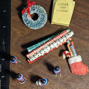 May include: Miniature Christmas-themed items on a brown surface. Includes wrapped gifts with blue bows, a green wreath with a red bow, a paper bag with text, rolled wrapping paper, toy soldiers, and a red stocking.