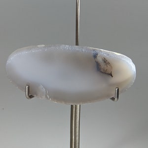 May include: A white agate slice with a dark gray inclusion, displayed on a clear acrylic stand with a metal rod.