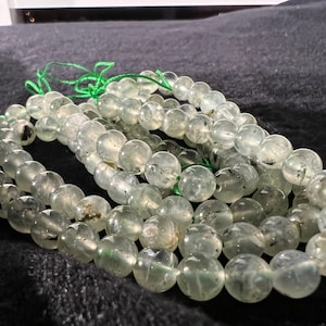 May include: A strand of light green gemstone beads with black inclusions. The beads are round and smooth, and they are strung on a green thread.