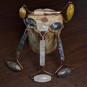 May include: A collection of facial massage rollers with copper-colored frames and various stone heads. The rollers feature green, blue, and clear quartz stones. The rollers are displayed on a wooden surface.