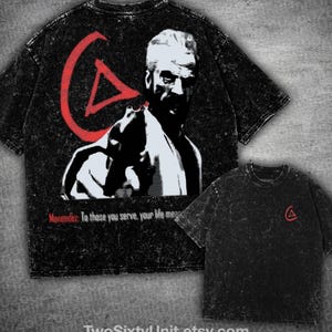 Puede incluir: Una camiseta negra con efecto lavado al ácido que presenta un gráfico grande de un hombre sosteniendo un arma de fuego, un símbolo de triángulo rojo y el texto "Menendez: To those you serve, your life means". El frente muestra un logo pequeño.