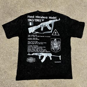 May include: Black t-shirt with a distressed finish, featuring a graphic of a firearm and technical specifications. The text "Pistol Mitralieră Model 1963/1965" is printed at the top. The shirt includes details about the rifle's mass, length, and firing rate.
