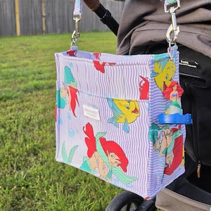 Op de afbeelding: Een kinderwagenorganisator met een paars en wit gestreept patroon met afbeeldingen van Ariel en Botje uit De Kleine Zeemeermin. De tas is met zilveren clips aan een kinderwagenhandvat bevestigd.