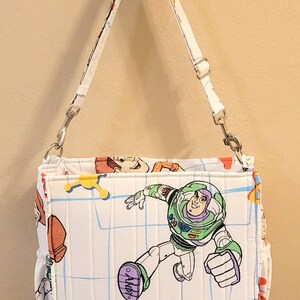 May include: White shoulder bag featuring a Buzz Lightyear graphic from Toy Story. The bag has a quilted pattern with a blue grid design and other character illustrations. It includes a white adjustable strap and silver-tone hardware.