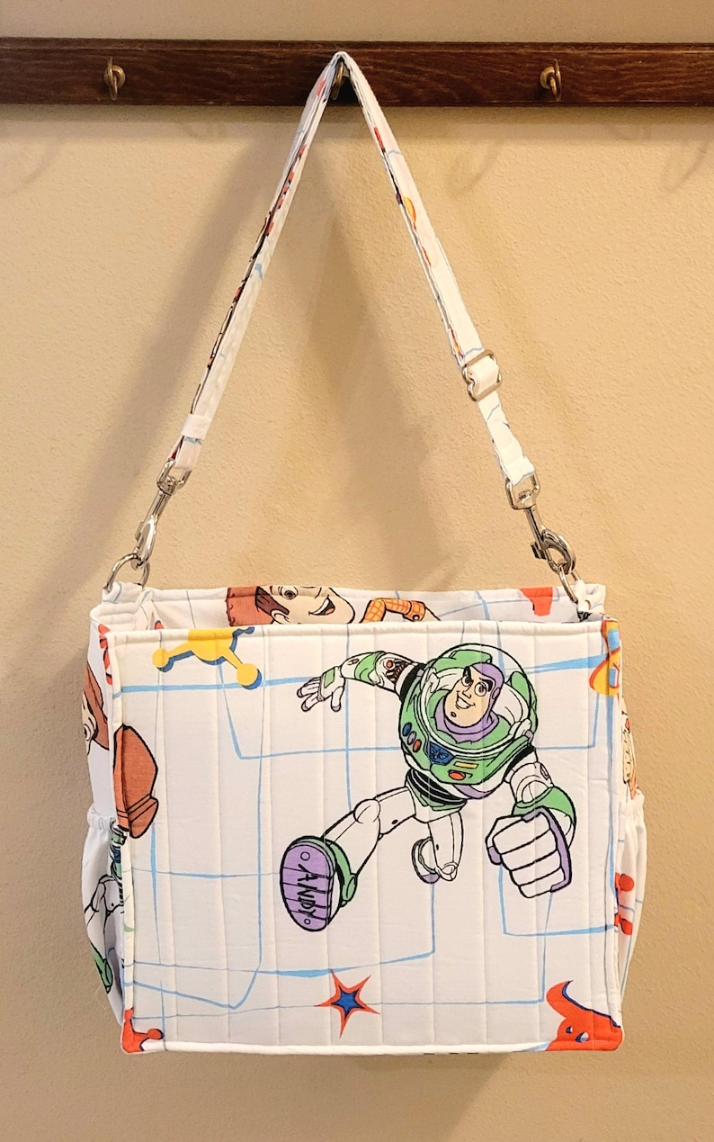 May include: White shoulder bag featuring a Buzz Lightyear graphic from Toy Story. The bag has a quilted pattern with a blue grid design and other character illustrations. It includes a white adjustable strap and silver-tone hardware.