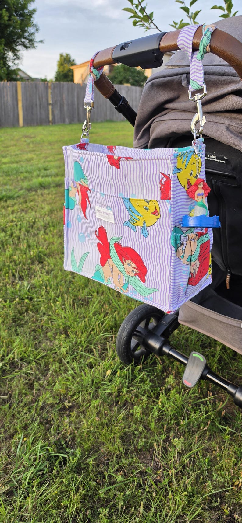Op de afbeelding: Een kinderwagenorganisator met een paars en wit gestreept patroon met afbeeldingen van Ariel en Botje uit De Kleine Zeemeermin. De tas is met zilveren clips aan een kinderwagenhandvat bevestigd.