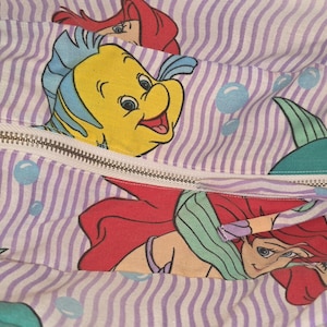 Op de afbeelding: Een stoffen tas met ritssluiting, met een herhalend patroon van verticale paarse strepen en cartoon illustraties van Ariel en Botje uit De Kleine Zeemeermin. De binnenkant van de tas is zichtbaar, met een paarse voering.