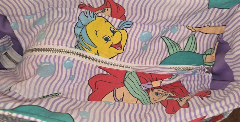 Op de afbeelding: Een stoffen tas met ritssluiting, met een herhalend patroon van verticale paarse strepen en cartoon illustraties van Ariel en Botje uit De Kleine Zeemeermin. De binnenkant van de tas is zichtbaar, met een paarse voering.