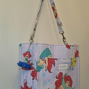 Op de afbeelding: Een draagtas met een herhalend patroon van een zeemeermin met rood haar en een blauwe staart, tegen een achtergrond van blauwe en witte verticale strepen. De tas heeft een bijpassende riem en een klein vak.