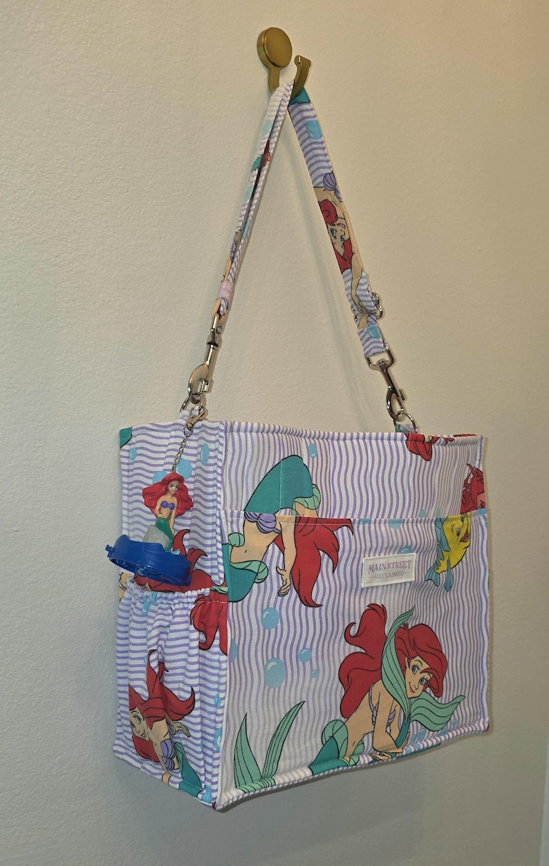 Op de afbeelding: Een draagtas met een herhalend patroon van een zeemeermin met rood haar en een blauwe staart, tegen een achtergrond van blauwe en witte verticale strepen. De tas heeft een bijpassende riem en een klein vak.