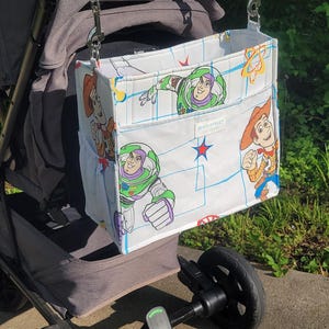 May include: A white stroller organizer bag featuring cartoon characters Buzz Lightyear and Woody. The bag is attached to a gray stroller with a brown handle. The bag has two pockets and is decorated with blue and red accents. The text "MAIN STREET" is visible.