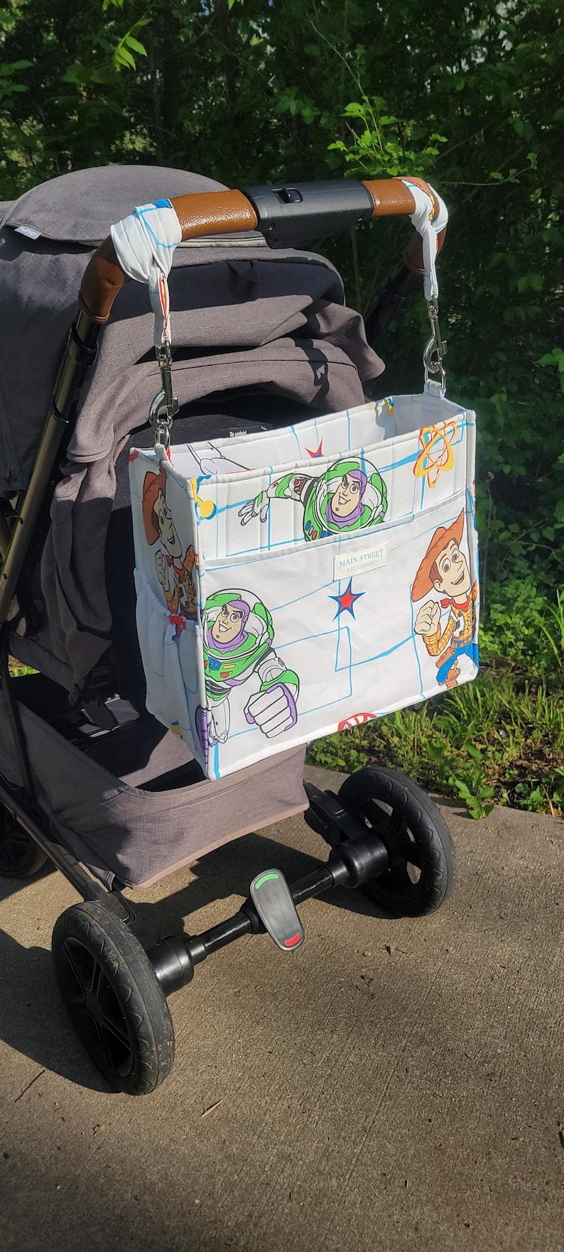 May include: A white stroller organizer bag featuring cartoon characters Buzz Lightyear and Woody. The bag is attached to a gray stroller with a brown handle. The bag has two pockets and is decorated with blue and red accents. The text "MAIN STREET" is visible.