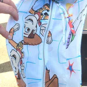 May include: White tote bag featuring a colorful print of characters from the animated film Toy Story. The bag has a zipper closure and a silver metal clasp. The interior is lined with the same print.