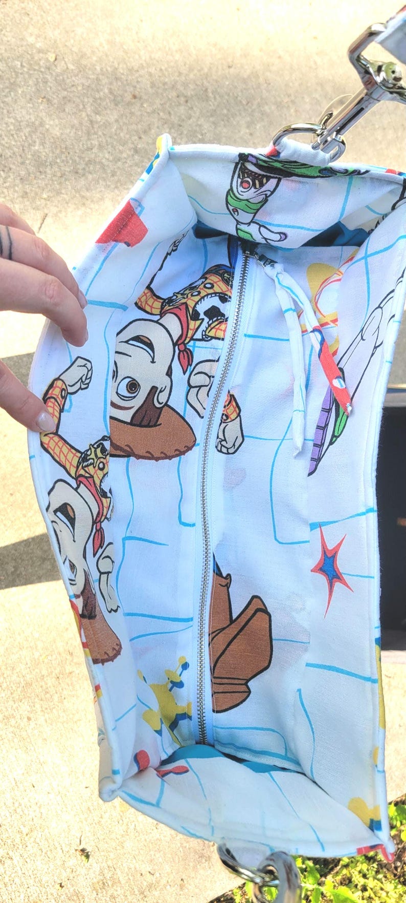 May include: White tote bag featuring a colorful print of characters from the animated film Toy Story. The bag has a zipper closure and a silver metal clasp. The interior is lined with the same print.