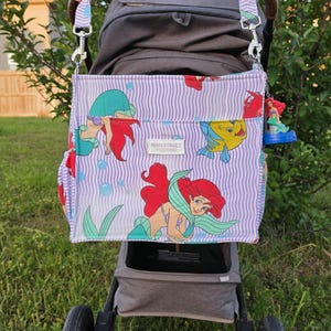 Op de afbeelding: Een kinderwagen met een paars en wit gestreepte luiertas met Ariel van De Kleine Zeemeermin. De tas heeft een bruin handvat en is bevestigd met zilveren clips. De kinderwagen staat op gras.