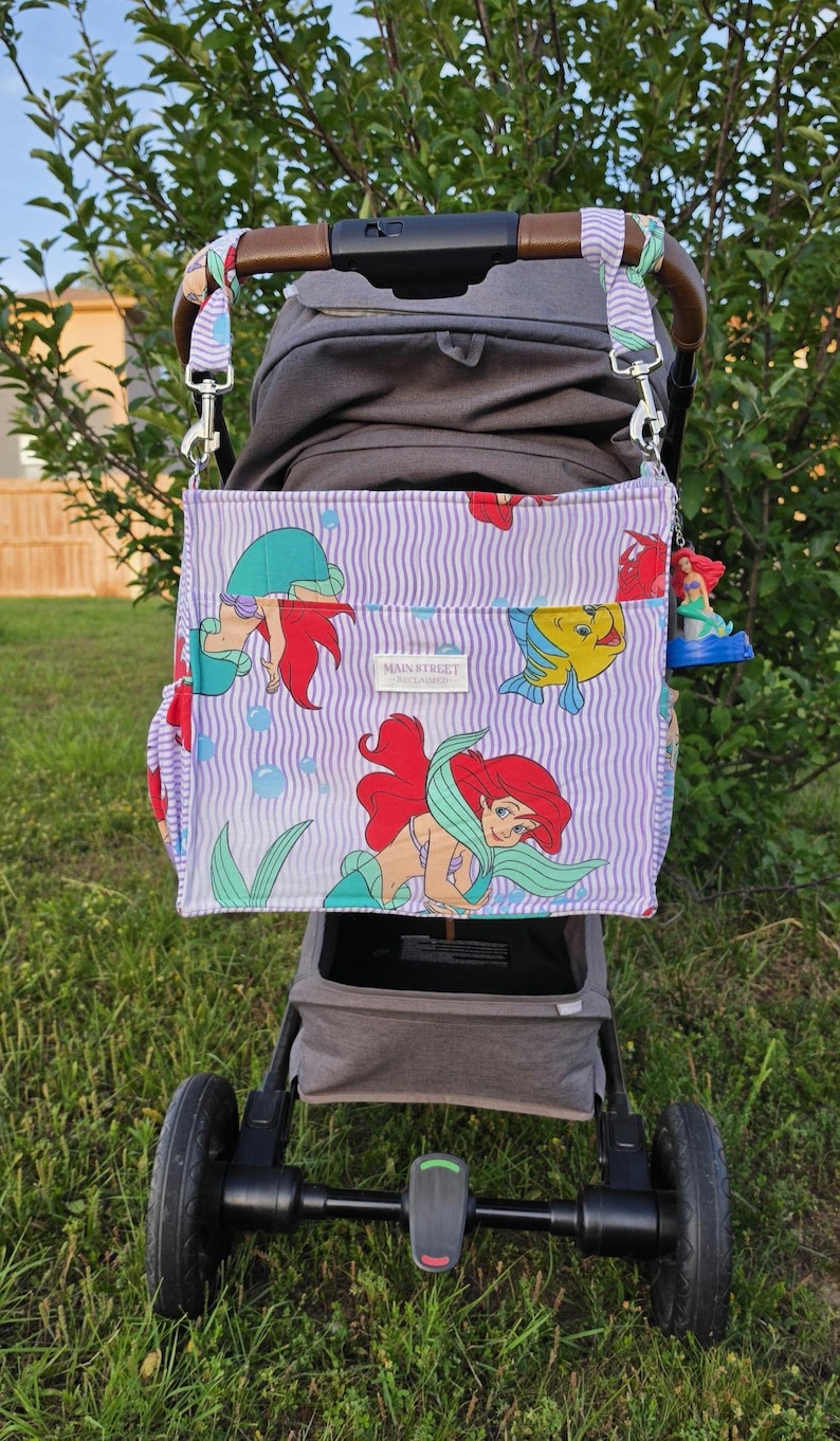 Op de afbeelding: Een kinderwagen met een paars en wit gestreepte luiertas met Ariel van De Kleine Zeemeermin. De tas heeft een bruin handvat en is bevestigd met zilveren clips. De kinderwagen staat op gras.