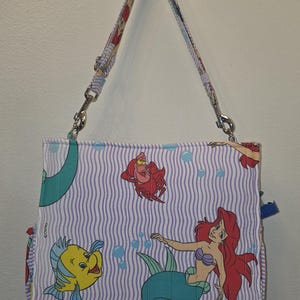 Op de afbeelding: Een rechthoekige handtas met een herhaald patroon van Ariel, Botje en Sebastiaan uit Disney's De Kleine Zeemeermin. De tas heeft een gestreepte achtergrond in wit en lichtpaars, met een bijpassende riem.