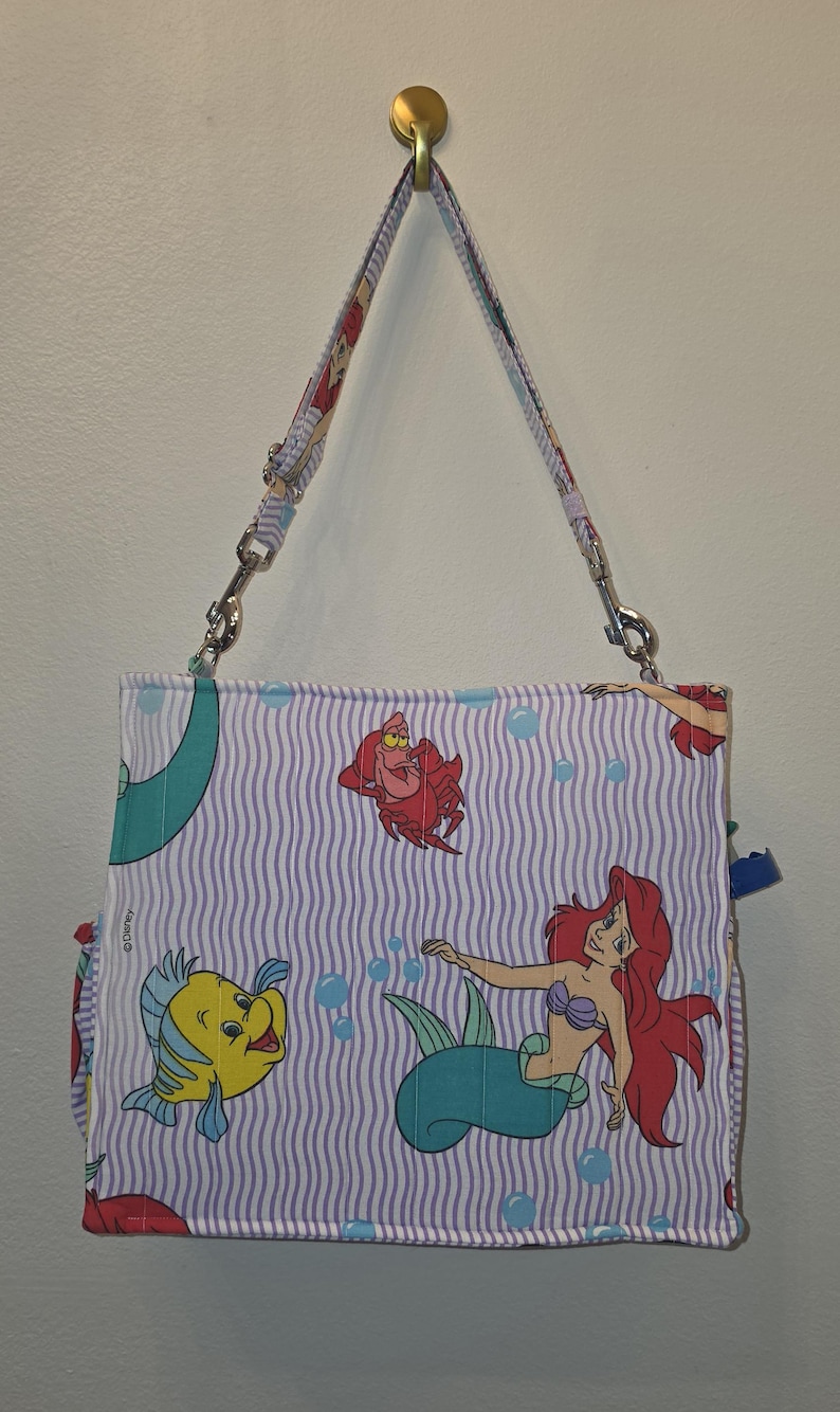 Op de afbeelding: Een rechthoekige handtas met een herhaald patroon van Ariel, Botje en Sebastiaan uit Disney's De Kleine Zeemeermin. De tas heeft een gestreepte achtergrond in wit en lichtpaars, met een bijpassende riem.