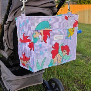 Op de afbeelding: Een kinderwagen organizer tas met een paars en wit gestreept patroon, met illustraties van een zeemeermin en vissen. De tas is bevestigd aan een grijze kinderwagen en heeft een bruine handgreep. De tas heeft een zijvak en de tekst "MAIN STREET" is zichtbaar.
