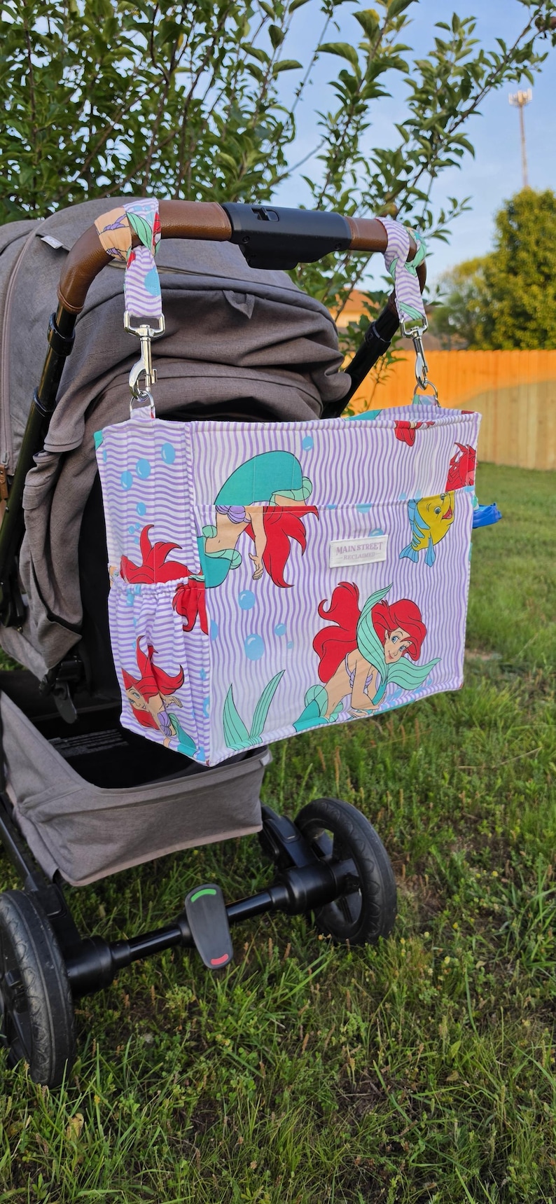 Op de afbeelding: Een kinderwagen organizer tas met een paars en wit gestreept patroon, met illustraties van een zeemeermin en vissen. De tas is bevestigd aan een grijze kinderwagen en heeft een bruine handgreep. De tas heeft een zijvak en de tekst "MAIN STREET" is zichtbaar.