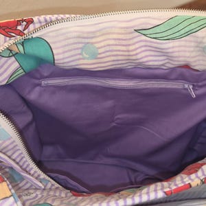 Op de afbeelding: Open tote bag met een zeemeerminthema. De tas heeft een paars en wit gestreept patroon met illustraties van zeemeerminnen en krabben. De binnenkant is effen paars en heeft een vak met ritssluiting. Een zilverkleurige metalen sluiting is aan de tas bevestigd.