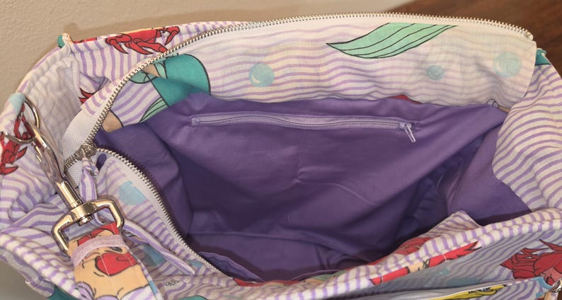 Op de afbeelding: Open tote bag met een zeemeerminthema. De tas heeft een paars en wit gestreept patroon met illustraties van zeemeerminnen en krabben. De binnenkant is effen paars en heeft een vak met ritssluiting. Een zilverkleurige metalen sluiting is aan de tas bevestigd.