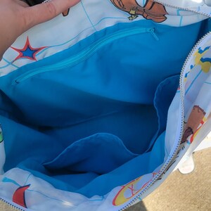May include: A white tote bag with a blue interior and a zipper closure. The exterior features a cartoon design with various colorful illustrations. The bag's interior includes a zippered pocket and additional compartments.