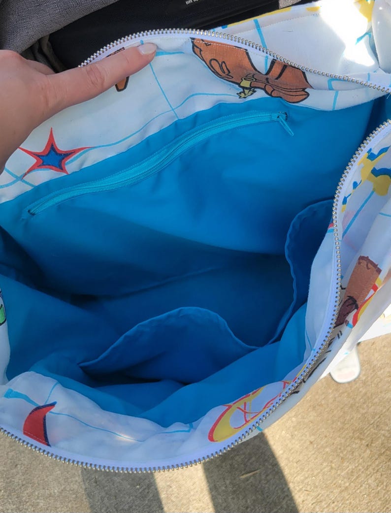 May include: A white tote bag with a blue interior and a zipper closure. The exterior features a cartoon design with various colorful illustrations. The bag's interior includes a zippered pocket and additional compartments.