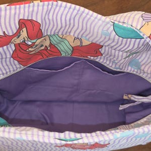 Op de afbeelding: Ariel-thema tote bag met een paarse binnenkant en een ritssluiting. De buitenkant heeft een herhalend patroon van Ariel, de Kleine Zeemeermin, op een wit en lavendel gestreepte achtergrond. Inclusief een zilverkleurige metalen sluiting.