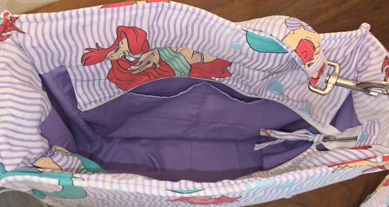 Op de afbeelding: Ariel-thema tote bag met een paarse binnenkant en een ritssluiting. De buitenkant heeft een herhalend patroon van Ariel, de Kleine Zeemeermin, op een wit en lavendel gestreepte achtergrond. Inclusief een zilverkleurige metalen sluiting.