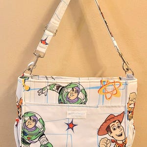 May include: White shoulder bag featuring Buzz Lightyear and Woody from Toy Story. The bag has a white strap and a front pocket with the text "MAIN STREET". The bag's design includes a blue grid pattern and cartoon graphics.