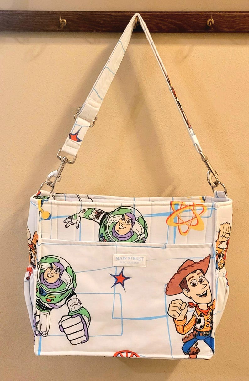 May include: White shoulder bag featuring Buzz Lightyear and Woody from Toy Story. The bag has a white strap and a front pocket with the text "MAIN STREET". The bag's design includes a blue grid pattern and cartoon graphics.