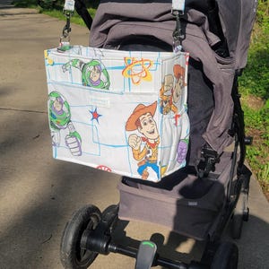 May include: A white stroller organizer bag featuring characters from the Toy Story movie. The bag is attached to a gray stroller with a brown handle. The bag has multiple pockets and a blue grid pattern.