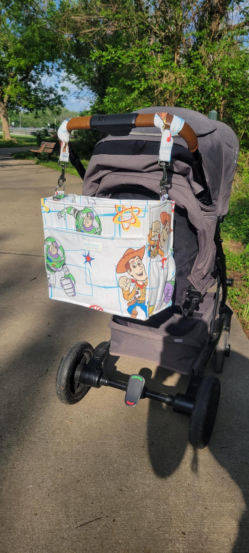 May include: A white stroller organizer bag featuring characters from the Toy Story movie. The bag is attached to a gray stroller with a brown handle. The bag has multiple pockets and a blue grid pattern.