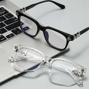 Puede incluir: Dos pares de gafas con lentes transparentes. Un par tiene una montura negra con detalles plateados, y el otro tiene una montura transparente con detalles plateados. Ambos pares tienen forma cuadrada y están sobre un portátil.