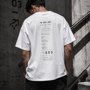May include: White t-shirt with black text on the back, including "VOID ARCHIVE UNIT 03" and other details. The shirt is made of 100% cotton. The person is wearing black cargo trousers. The background is a concrete wall with the number "03" on it.