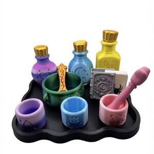 May include: A collection of miniature potion-making accessories on a black scalloped tray, including three colorful bottles with gold caps, a green cauldron with a gold spoon, a gray book, and three small cups in purple, blue, and pink.