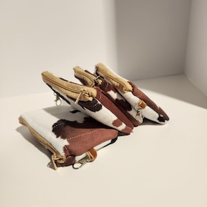 May include: Three rectangular pouches featuring a brown and white cow print pattern. Each pouch has a tan zipper closure along the top edge and a small metal lobster clasp attached to one side, allowing for easy attachment to other items.