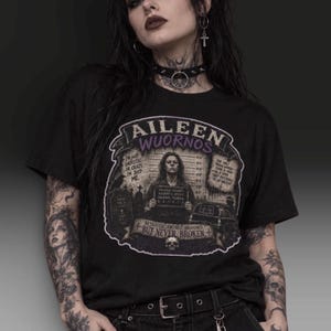 May include: A black short-sleeved t-shirt featuring a graphic print of Aileen Wuornos. The design includes a mugshot, a tombstone, a car, and the text: Aileen Wuornos, I'm not sadistic or crazy, I'm just me, Betrayed, Abused, Branded, But Never Broken.