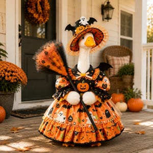 May include: A white goose figurine dressed in a Halloween-themed outfit featuring an orange and black dress with pumpkin, ghost, and bat patterns. The goose wears a matching wide-brimmed hat and holds a small broom with orange and black bristles.