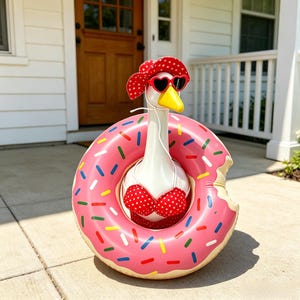 May include: A white goose figurine wearing a red and white polka dot sun hat, heart-shaped sunglasses, and a matching red and white polka dot bikini top, positioned inside a large, pink, inflatable donut pool float with colorful sprinkles.