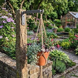 May include: A hanging terracotta pot filled with greenery and flowers, suspended by a macrame plant hanger from a metal bracket attached to a stone pillar. The garden setting features a variety of plants and a greenhouse in the background.