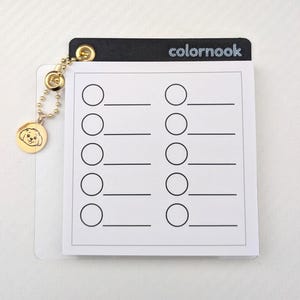 以下が含まれることがあります： 銀色のグリッターで「colornook」の文字が入った黒いトップの白いメモ帳。メモ帳には、下に線が入った10個の円があります。犬の顔の金色のチャームが金のチェーンからぶら下がっています。